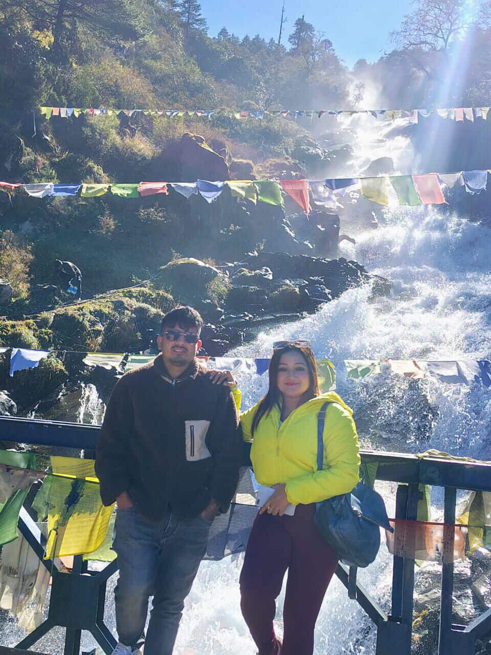 A honeymoon couple standing close together beside a misty Himalayan waterfall on a Sikkim road trip, green forest and wet rocks around them.