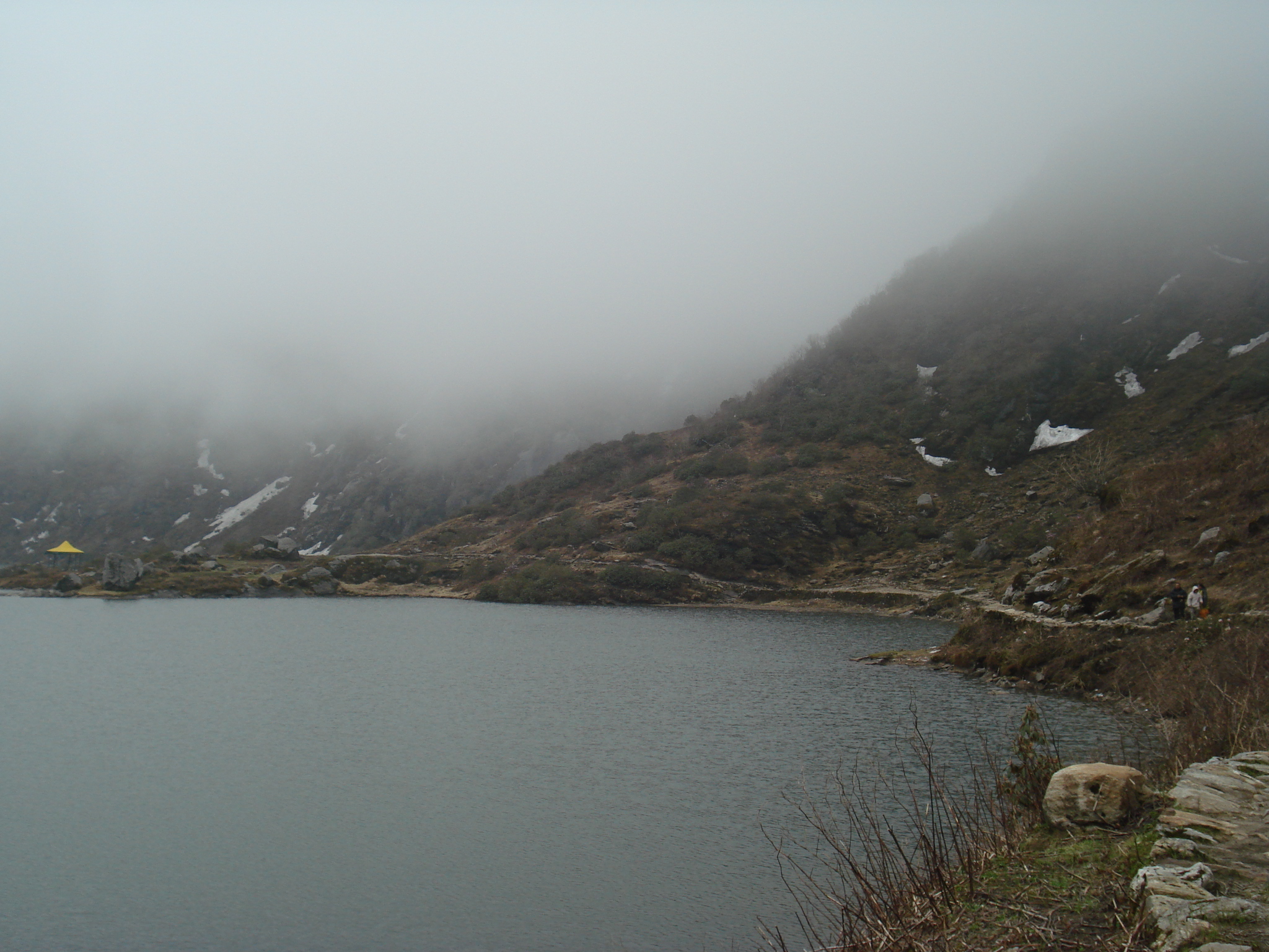 Turquoise glacial waters of Tsomgo (Changu) Lake in East Sikkim ringed by snow-dusted mountains under a clear blue sky, with prayer flags on the shore.