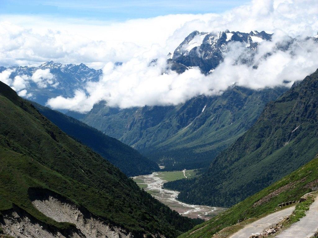 The wide green meadow of Yumthang Valley in North Sikkim in spring, with a river winding through and snow-dusted Himalayan peaks rising on either side.